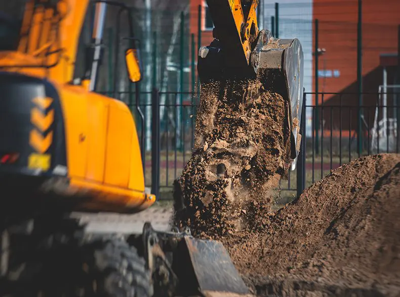 Excavator scooping and dumping dirt at a construction site, with a blurred background of fencing and buildings.